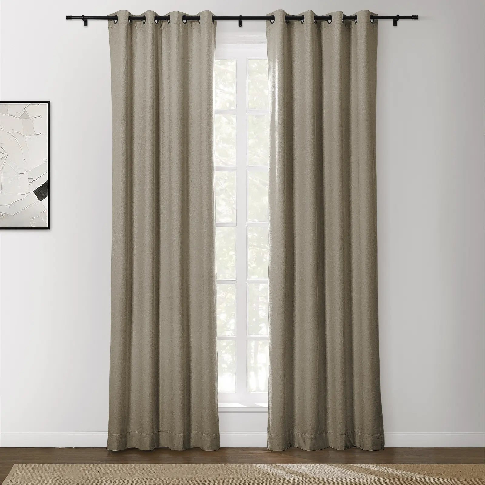 A pair of long greige grommet blackout curtains on a black rod in a modern room with a bright window, an abstract white art print, and a woven beige rug on a dark wood floor
