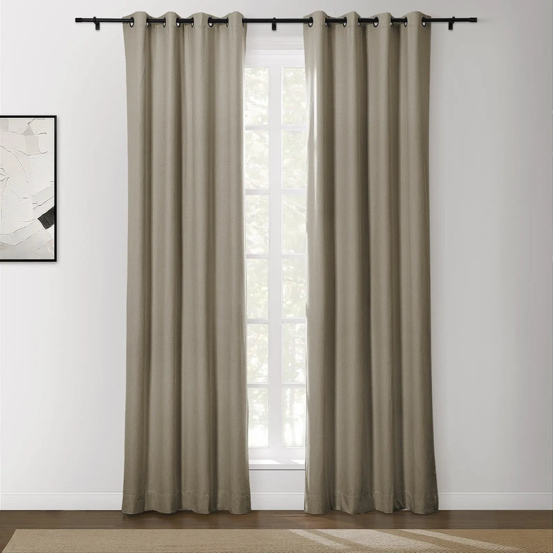 A pair of long greige grommet blackout curtains on a black rod in a modern room with a bright window, an abstract white art print, and a woven beige rug on a dark wood floor