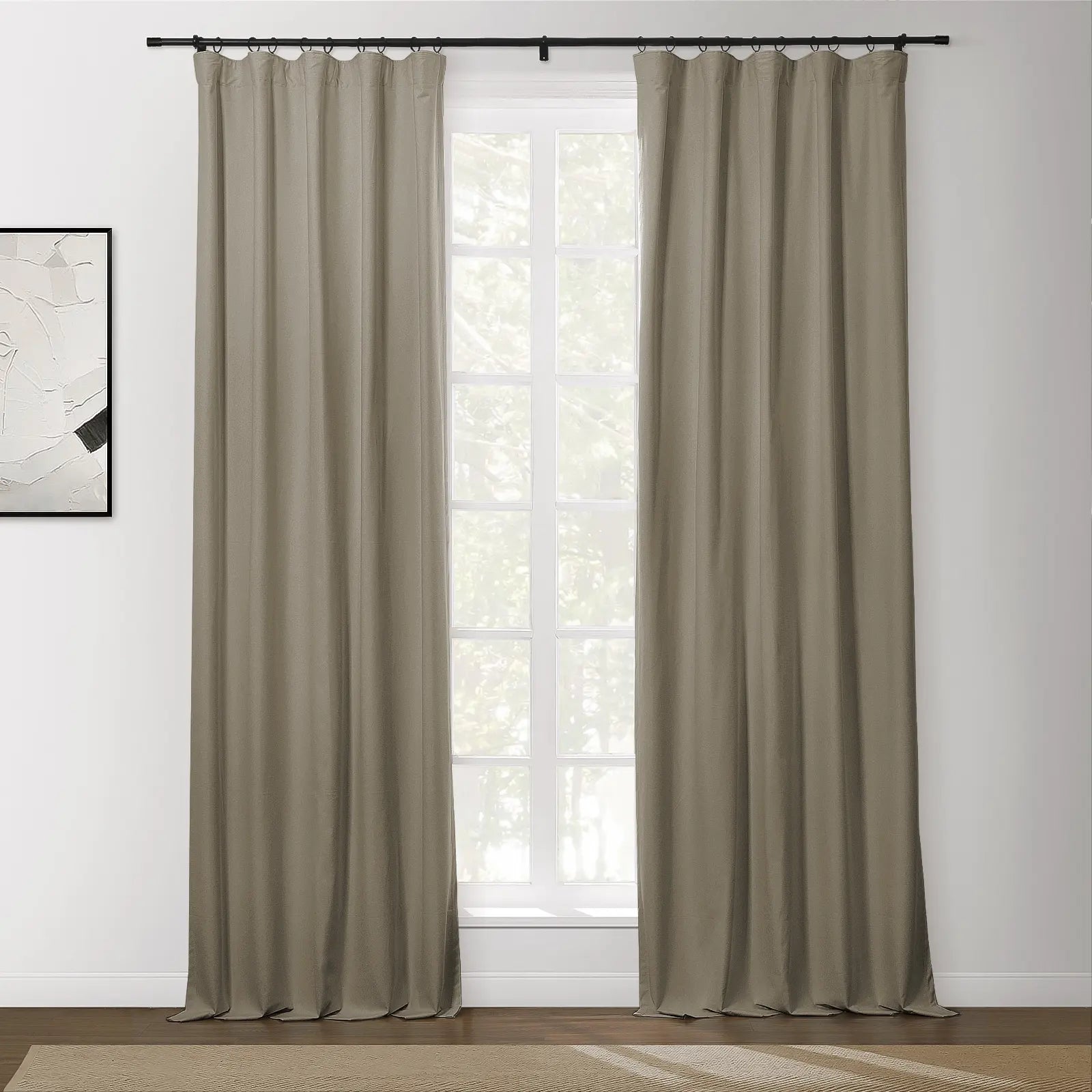 A pair of taupe grommet curtains on a black rod flanking a bright multi-paned window in a modern room with abstract wall art and a beige textured rug