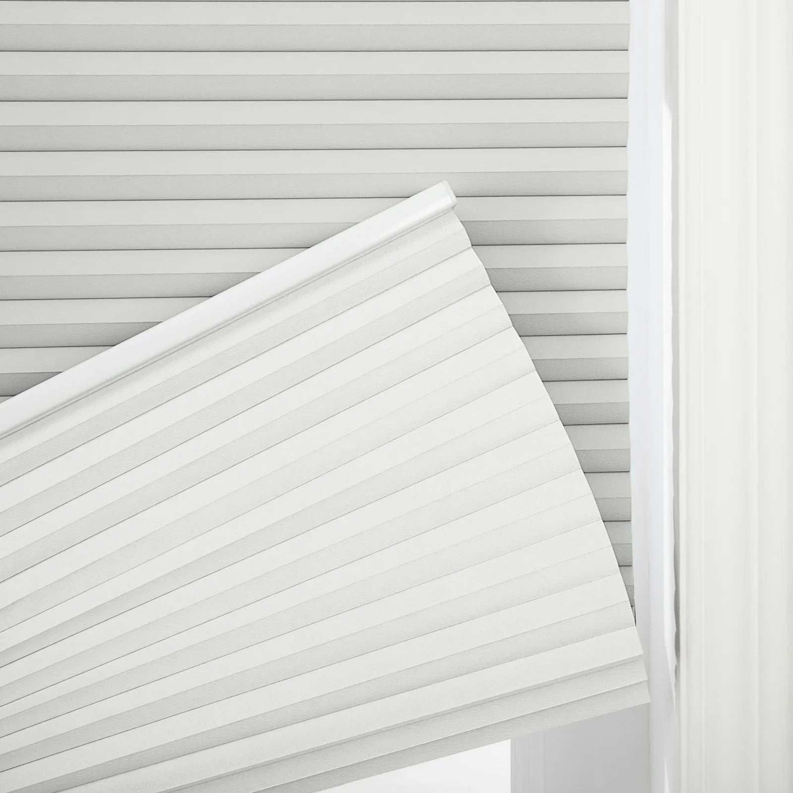 A bright white cellular window Blind is partially raised displaying crisp uniform accordion pleats against a subtle matching pleated background and a soft sheer curtain edge to the right
