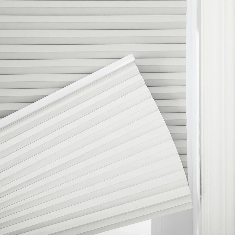 A close-up of a partially rolled light grey cellular pleated window Blind revealing its accordion-like structure, next to a soft white sheer curtain