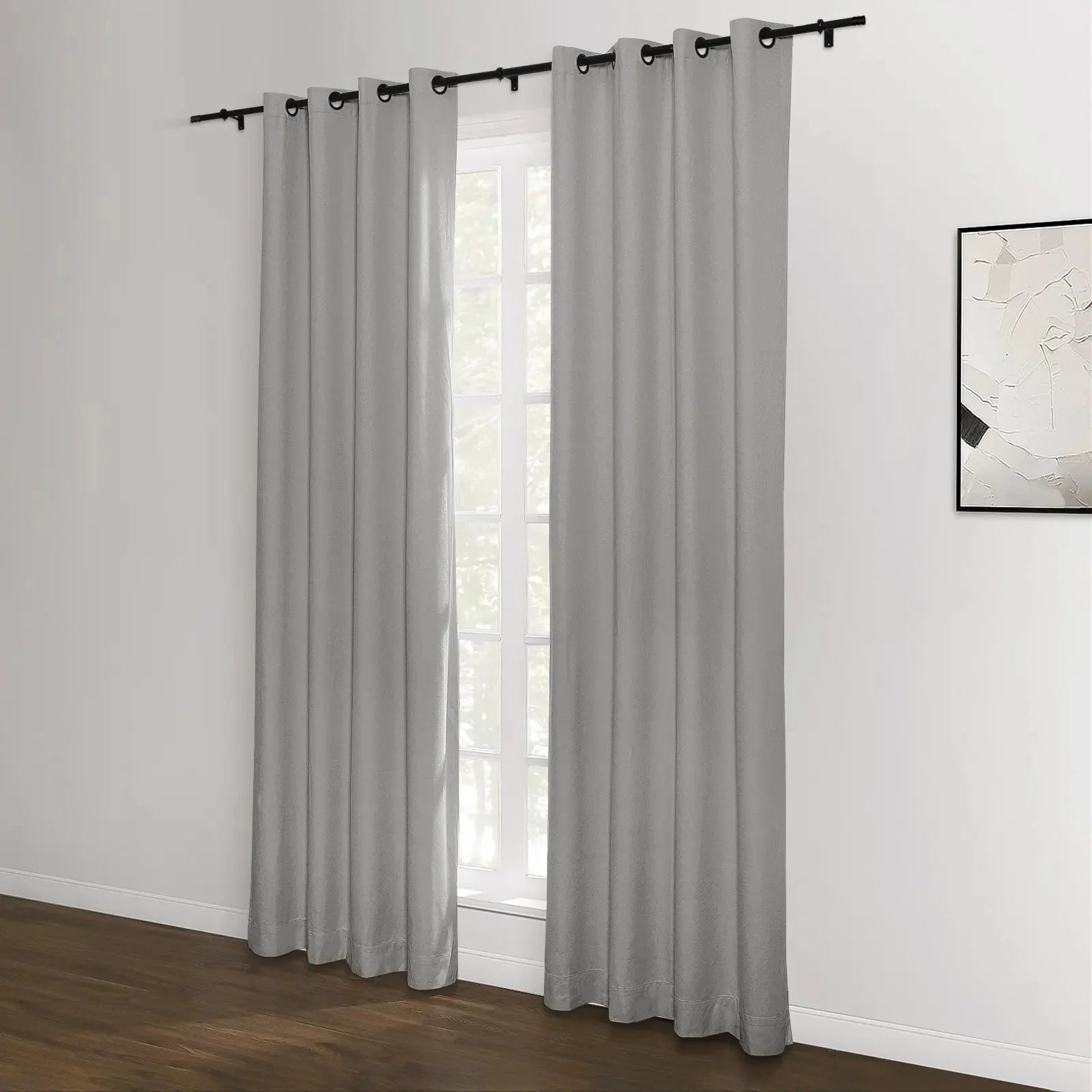 Two grey grommet top curtains on a black rod frame a window with natural light in a room with dark wood flooring and a white wall displaying an abstract black and white framed artwork
