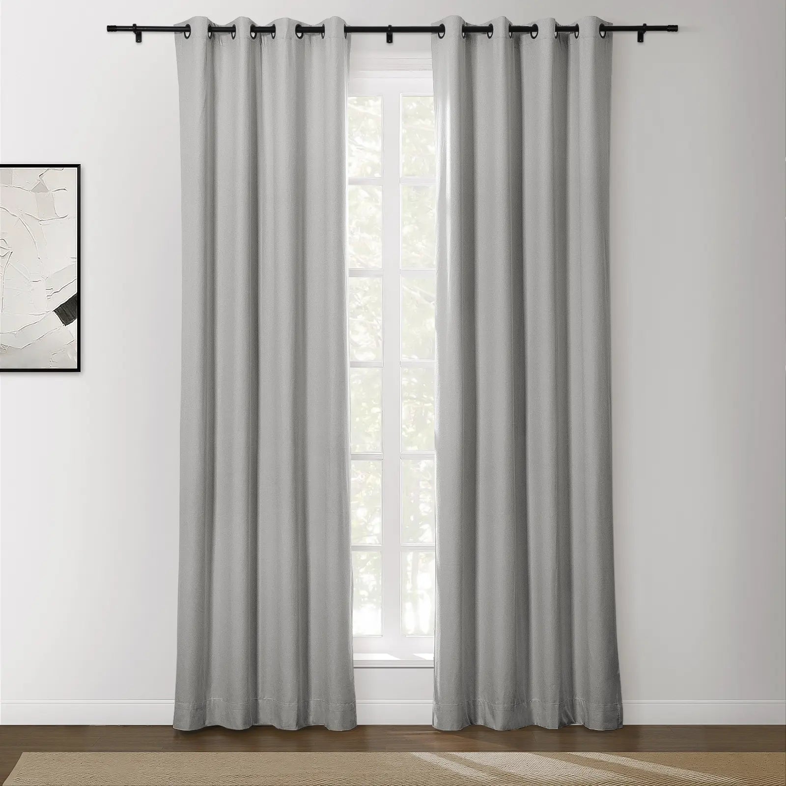 Photograph of light gray grommet top curtains on a black rod framing a window in a bright room, with an abstract white and black artwork on the wall and a brown textured rug on the wooden floor