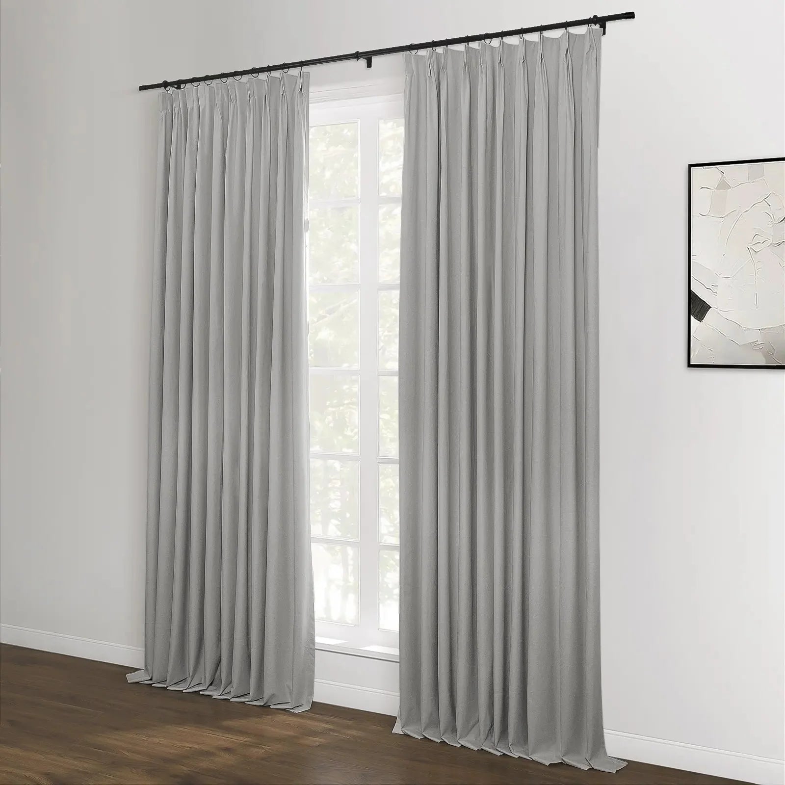 Photograph of light gray pleated curtains on a black rod over a large window showing green trees, with a textured white abstract artwork on the adjacent white wall