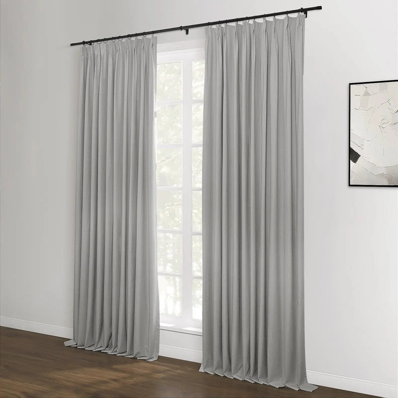 Photograph of light gray pleated curtains on a black rod over a large window showing green trees, with a textured white abstract artwork on the adjacent white wall