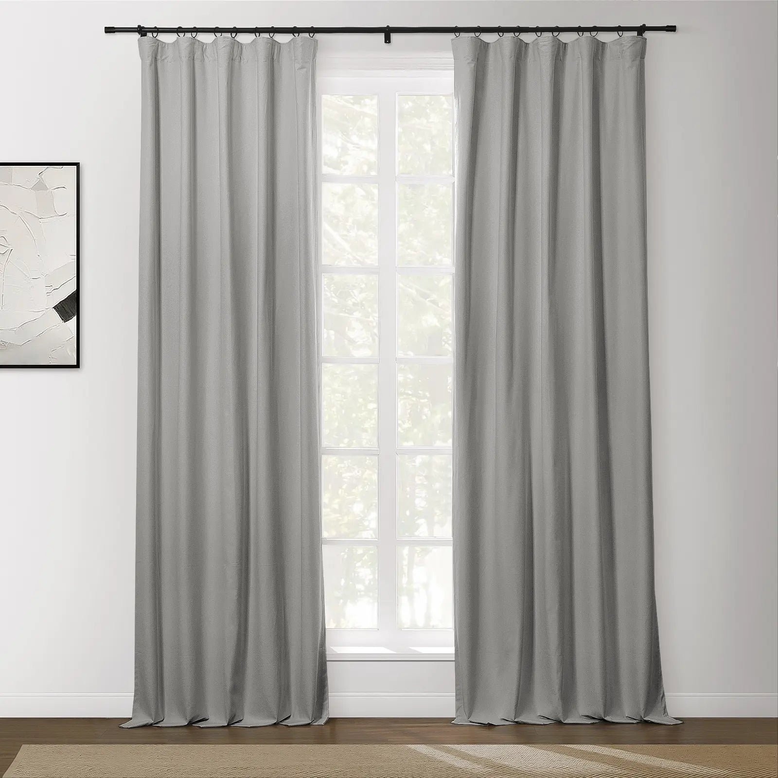 Light grey panel curtains hang from a black rod with rings covering a white-framed window in a room with an abstract white and black artwork and a textured brown rug