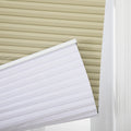 Photograph of a beige cellular Blind fully closed at the top of a window, with a white pleated Blind partially lowered at a diagonal angle showing its crisp texture