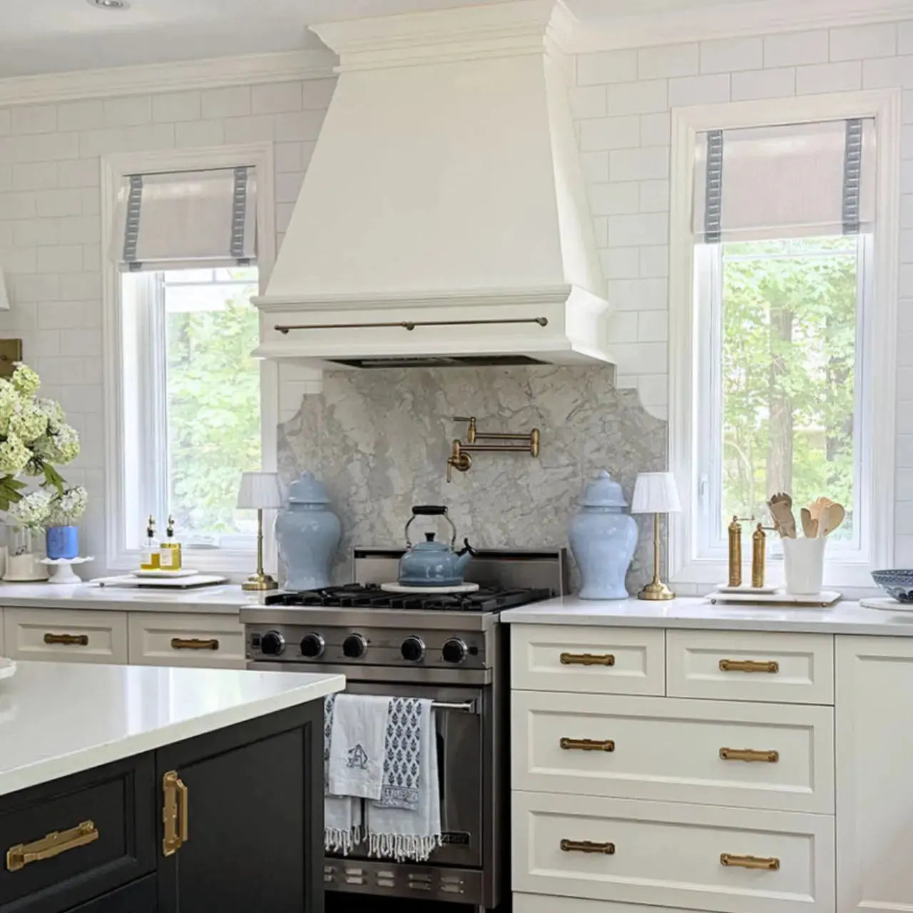 A bright modern kitchen with a large white range hood above a marble backsplash, a stainless steel range topped with a blue teapot, and pale blue ginger jars accenting white and dark cabinets with gold hardware