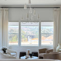 A stylish modern living room with a neutral palette showcasing a unique white multi-cone chandelier, elegant cream drapes, and a mix of beige velvet and white boucle armchairs, overlooking distant verdant hills and a construction site