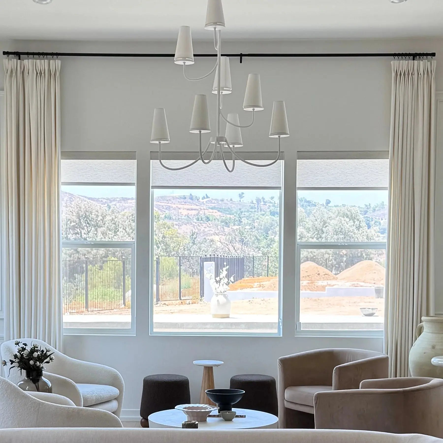 A stylish modern living room with a neutral palette showcasing a unique white multi-cone chandelier, elegant cream drapes, and a mix of beige velvet and white boucle armchairs, overlooking distant verdant hills and a construction site