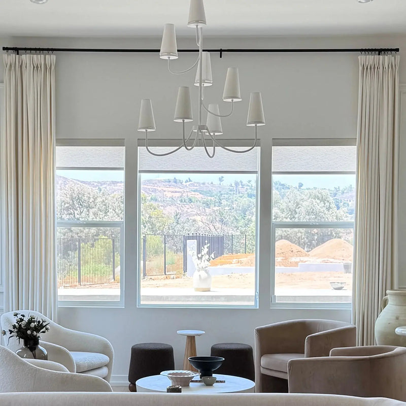 A stylish modern living room with a neutral palette showcasing a unique white multi-cone chandelier, elegant cream drapes, and a mix of beige velvet and white boucle armchairs, overlooking distant verdant hills and a construction site