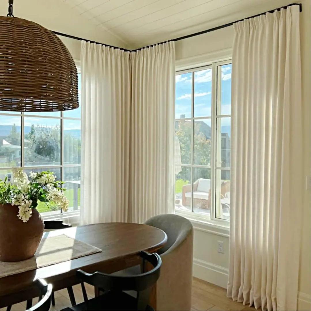 Photograph of an elegant dining room with a large woven wicker pendant light, white pleated curtains on black rods, a dark wood dining table holding a rustic pot of white flowers, and a serene window view of distant green hills under a blue sky