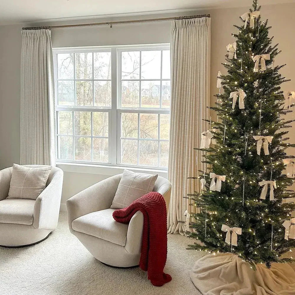 A modern living room decorated for Christmas featuring a large green Christmas tree adorned with white bows and warm string lights, two cream-colored upholstered swivel chairs, one with a vibrant red chunky knit throw blanket, all positioned before a multi-paned window looking out to a wintery landscape