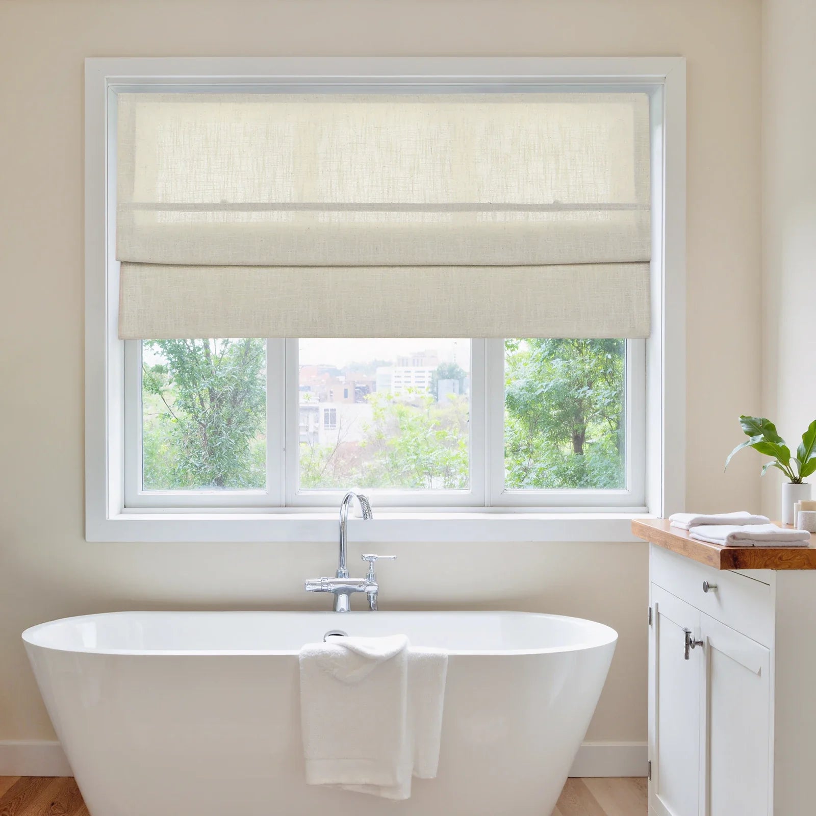 Photograph of a serene bathroom featuring a freestanding white tub with a chrome faucet, beige linen Roman shades over a window revealing a lush green outdoor view with distant city buildings, and a small white vanity with a wooden top