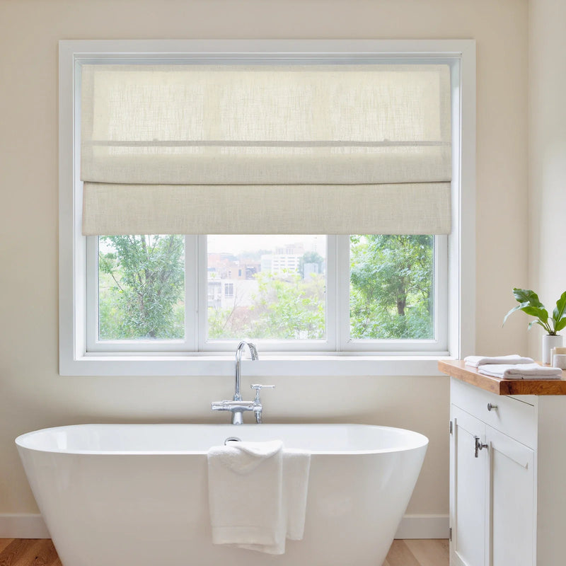 Photograph of a serene bathroom featuring a freestanding white tub with a chrome faucet, beige linen Roman shades over a window revealing a lush green outdoor view with distant city buildings, and a small white vanity with a wooden top