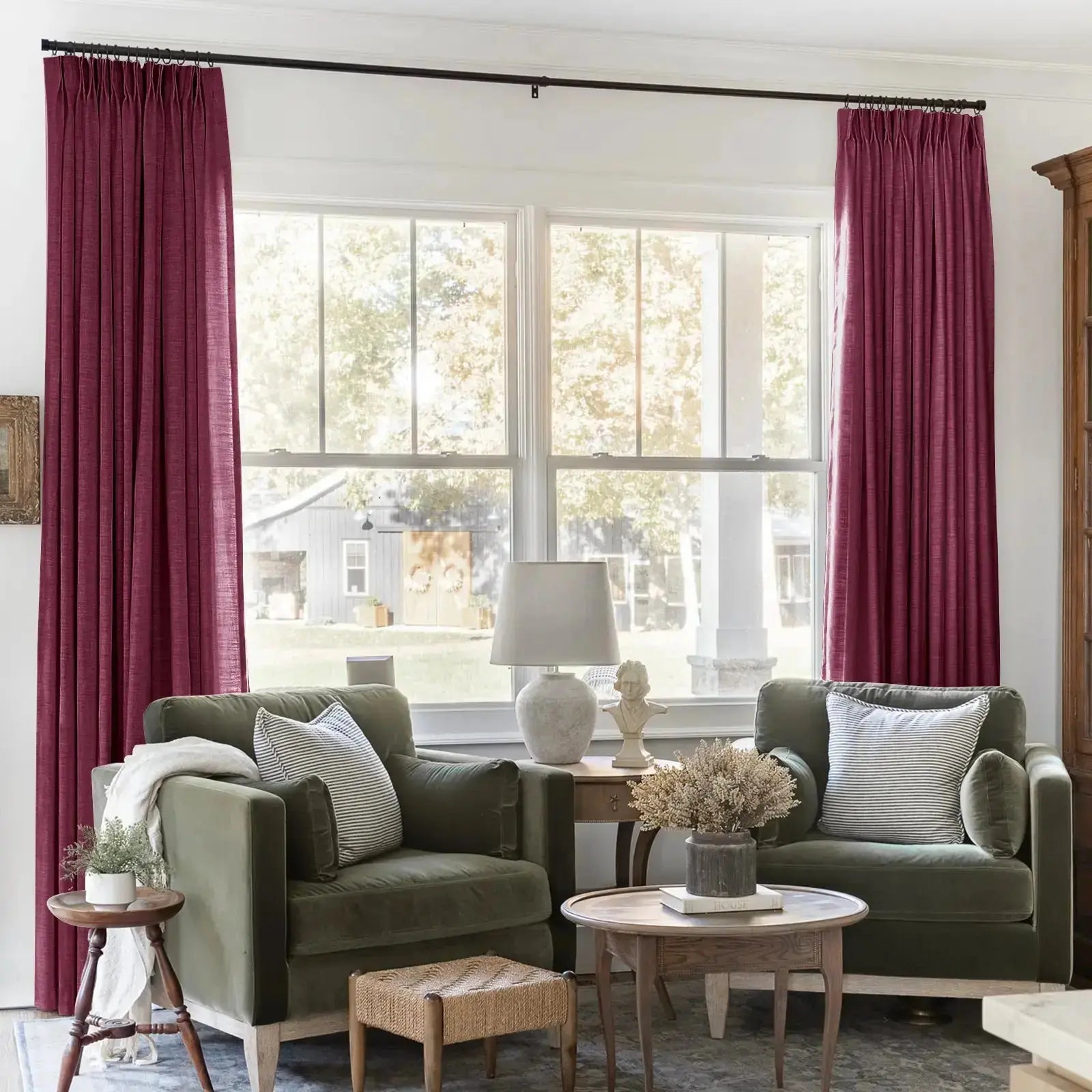 Cozy living room with olive-green velvet armchairs and striped cushions, magenta pinch-pleat curtains framing a large window overlooking a rustic barn, featuring a white classical bust and textured ceramic lamp