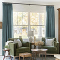 A rustic living room interior featuring two olive green velvet armchairs, striped throw pillows, blue drapes, and a large window overlooking a grey barn with two decorative wreaths and a white table lamp with a bust statue
