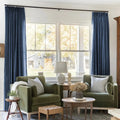 Interior photograph of a cozy living room with two olive green velvet armchairs flanking a large window offering a view of a white barn with wreaths outside, highlighted by dark blue pleated curtains and a decorative bust sculpture