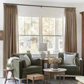 A bright living room with two olive green velvet armchairs, striped pillows, and warm brown curtains framing a window overlooking a rustic barn and white porch column, featuring a ceramic lamp, classical bust, and dried flower arrangement