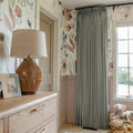 A cozy nursery corner featuring watercolor floral wallpaper in blush, blue, and brown hues, a woven rattan table lamp on a light wood dresser, and sage green curtains next to a window with a green outdoor view and a wicker basket of stuffed animals