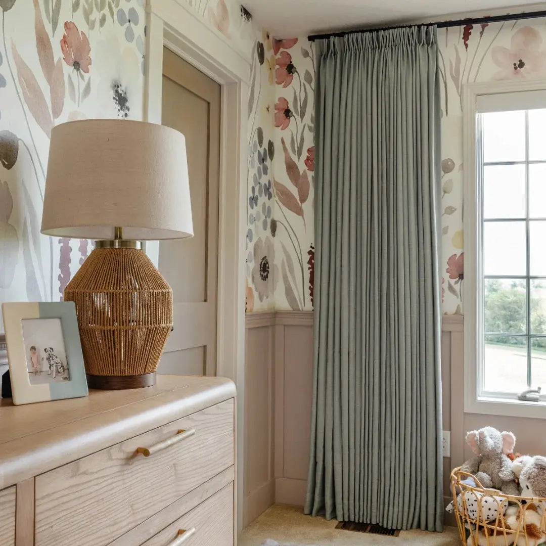 A cozy nursery corner featuring watercolor floral wallpaper in blush, blue, and brown hues, a woven rattan table lamp on a light wood dresser, and sage green curtains next to a window with a green outdoor view and a wicker basket of stuffed animals