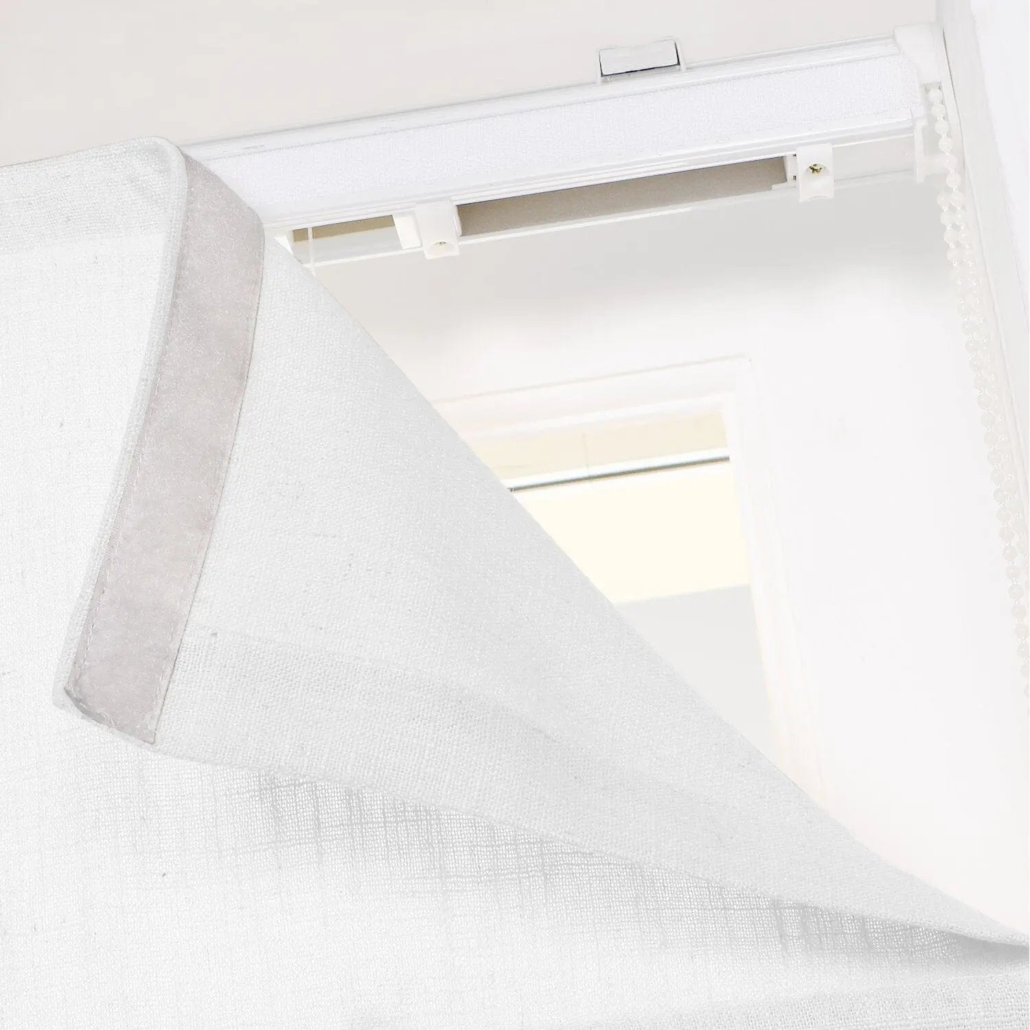 Close-up of a white textured fabric window blind or Roman shade panel showing the Velcro attachment to a top track system with a white beaded chain operator indoors