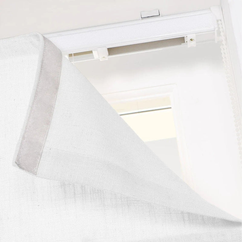Close-up of a white textured fabric window blind or Roman shade panel showing the Velcro attachment to a top track system with a white beaded chain operator indoors