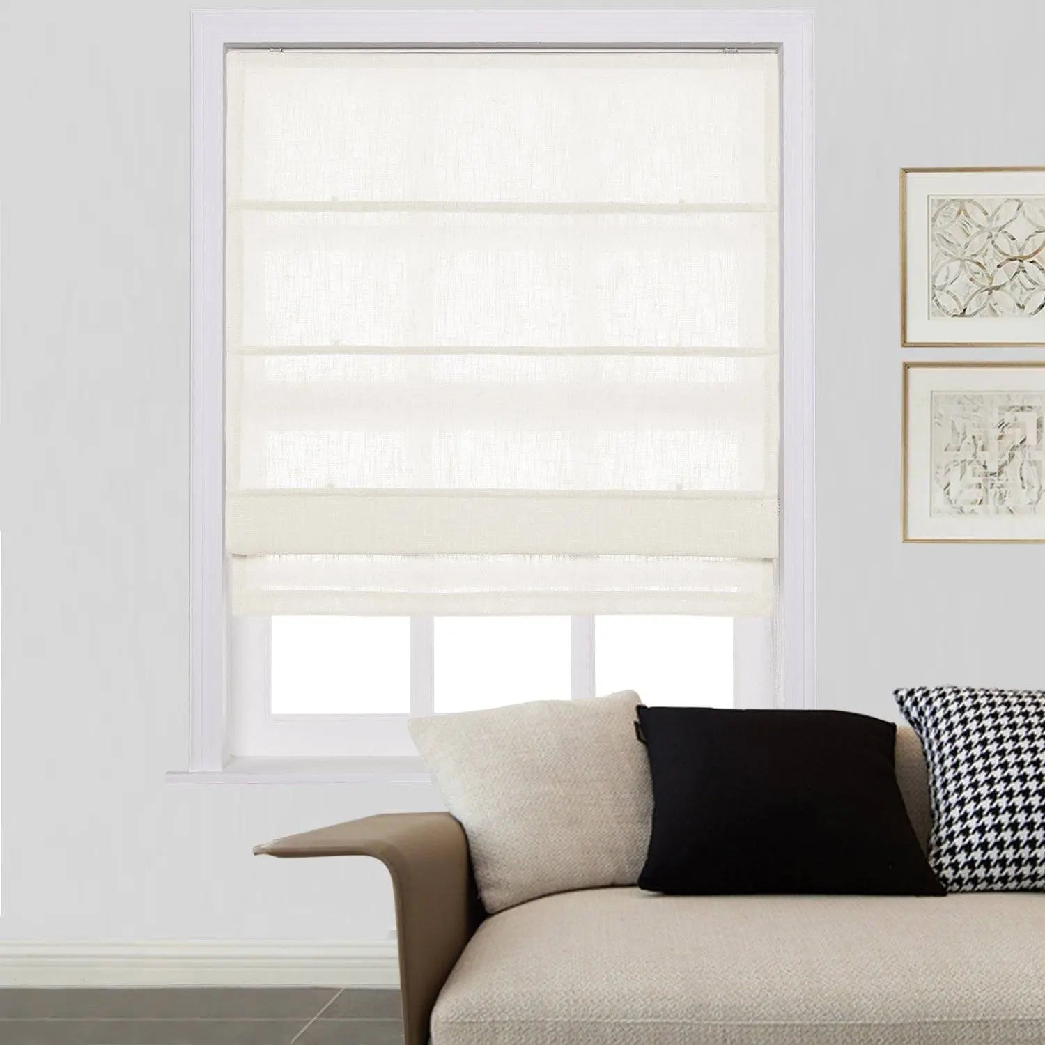 Photograph of a modern interior featuring a window with a white linen Roman blind, a light beige sofa with black and houndstooth patterned pillows, and two framed abstract artworks