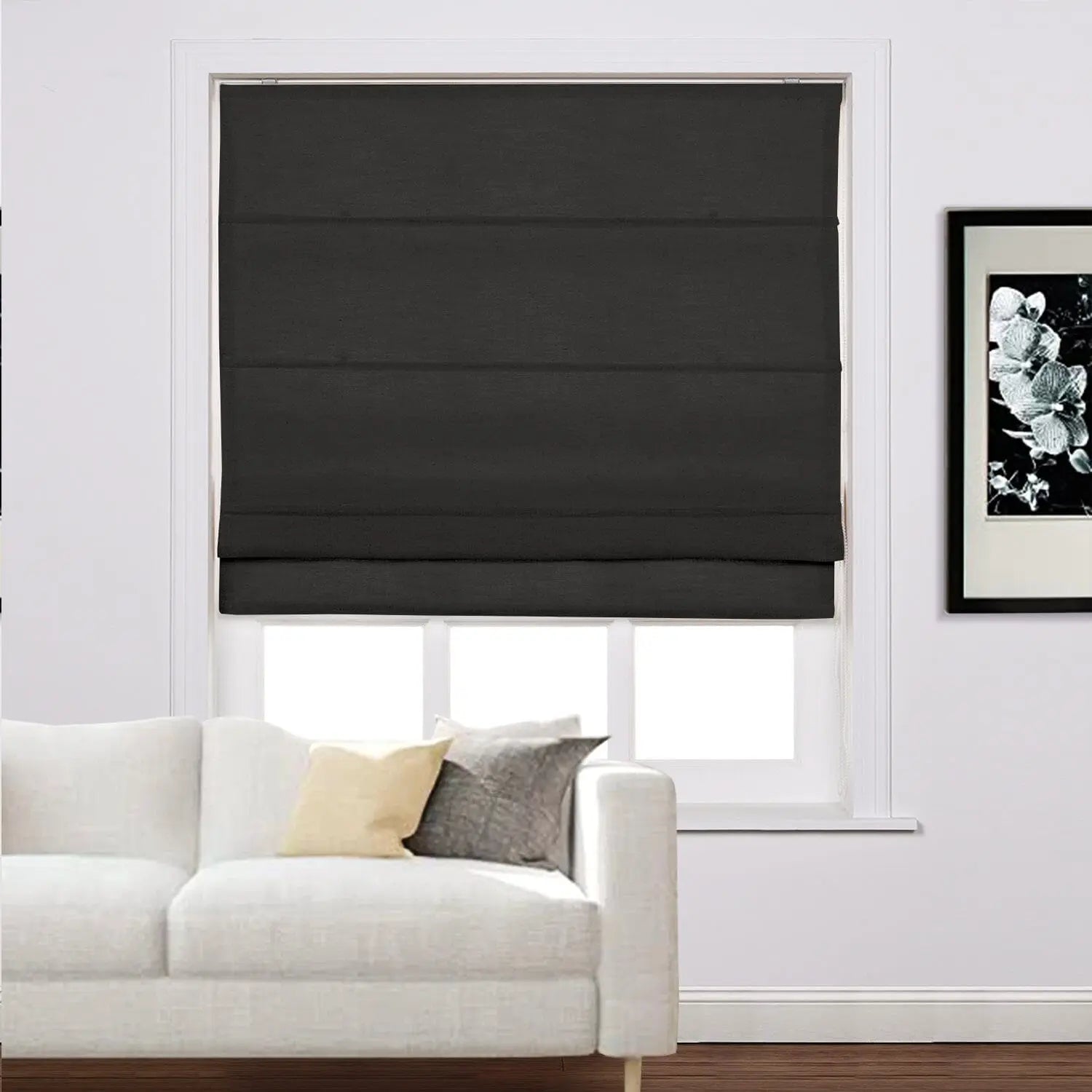 Black fabric roman shade covering a window in a modern living room featuring a white sofa with light yellow and charcoal grey throw pillows and a black framed floral art piece