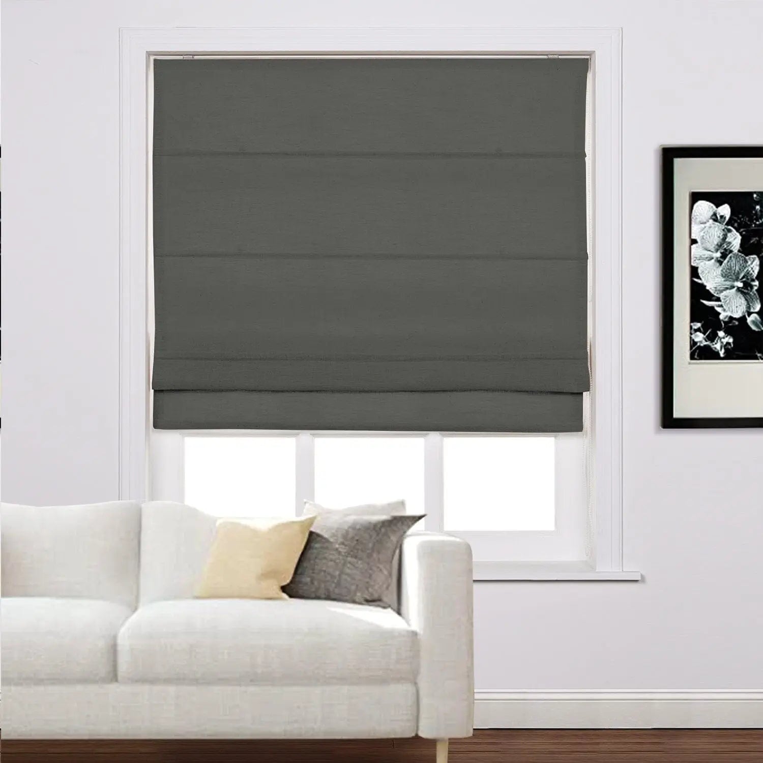 A modern living room showcasing a dark grey fabric Roman blind on a window, a white minimalist sofa accented with beige and grey pillows, and black-framed white floral artwork