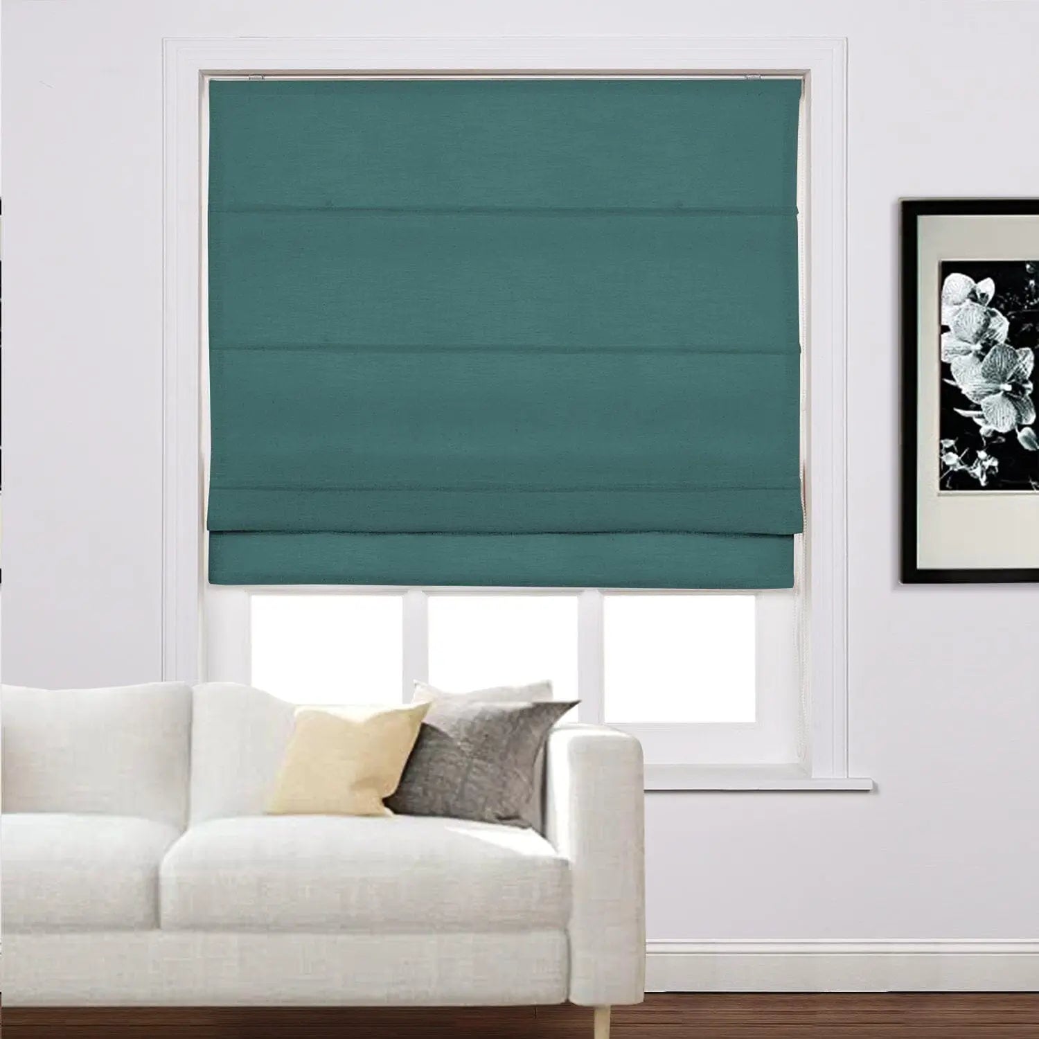 A home interior featuring a window with a teal Roman blind above a white sectional sofa adorned with light yellow and grey pillows and a black and white floral art print on the wall