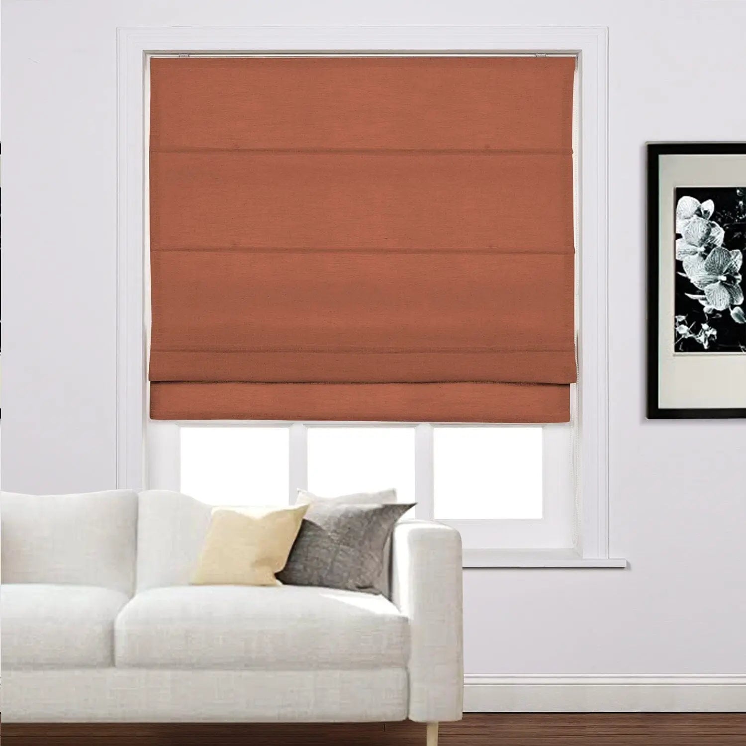 Photograph of a modern living room interior featuring a warm terracotta Roman shade on a white window frame, a plush white tufted sofa with cream and grey throw pillows, and a framed black and white orchid artwork