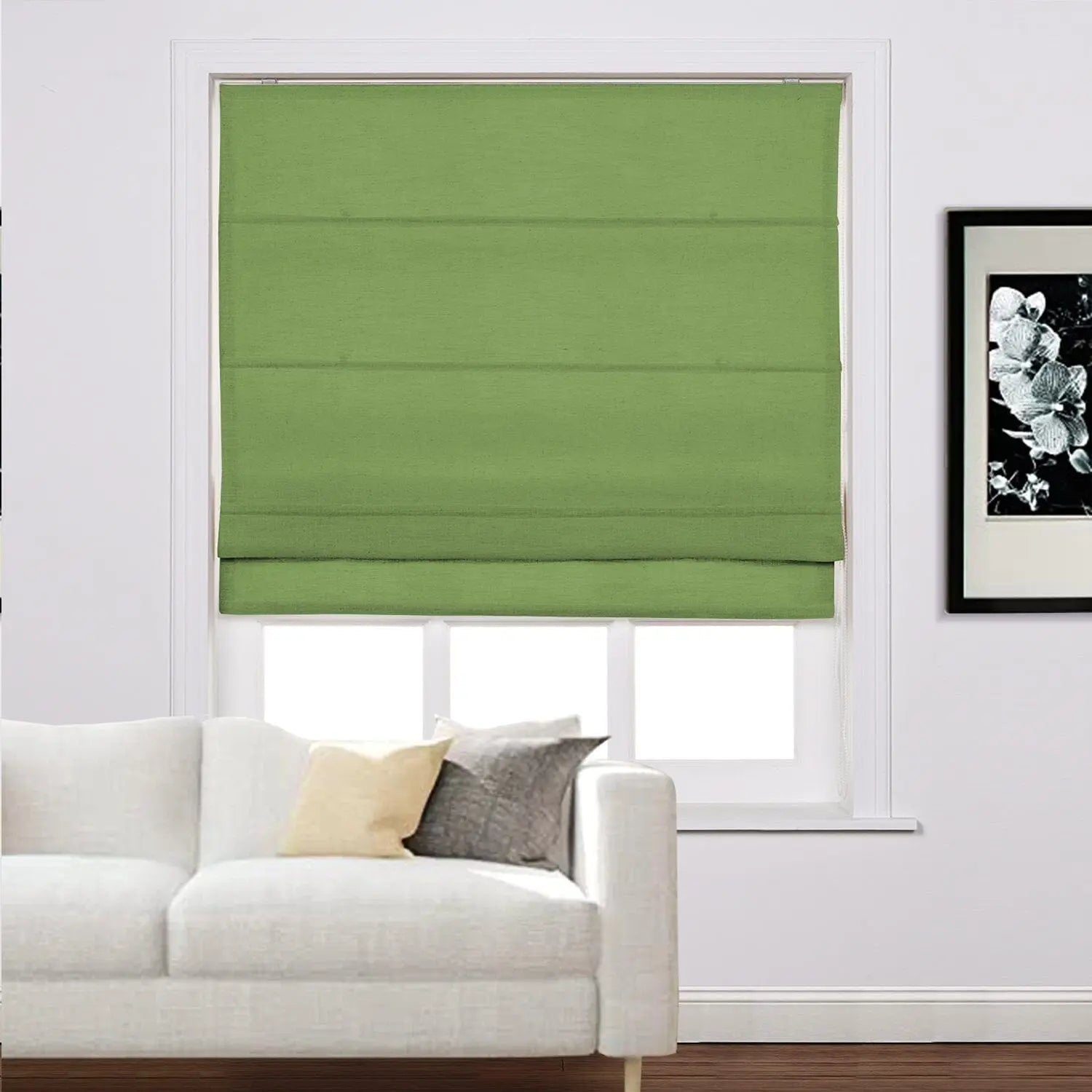 A green Roman blind partially covers a window in a contemporary living room featuring a white sofa with beige and gray pillows and framed black and white floral artwork