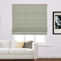 A modern living room interior with a window featuring a textured sage green Roman blind, a plush white sofa with decorative pillows, and a framed black and white floral artwork on the wall