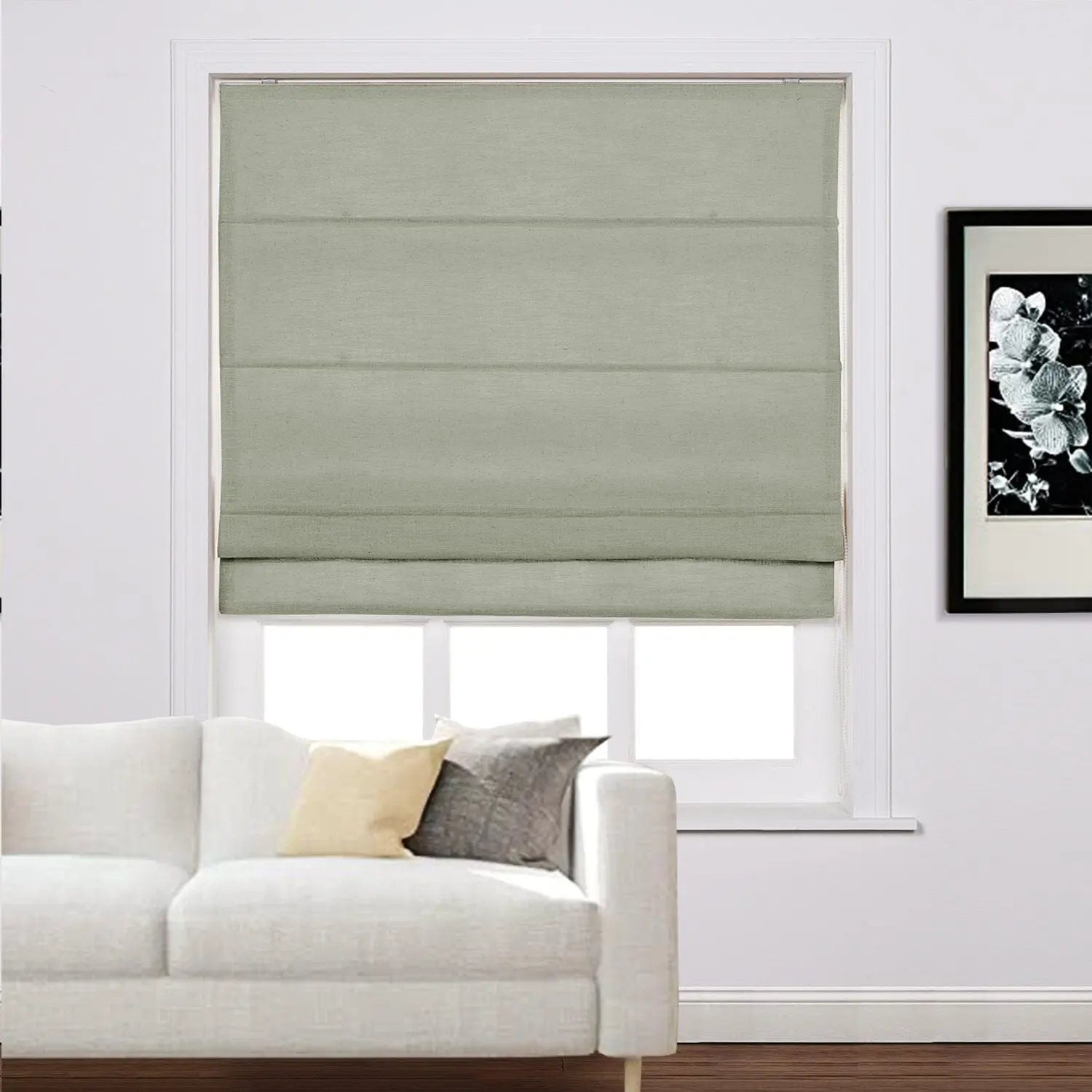 A modern living room interior with a window featuring a textured sage green Roman blind, a plush white sofa with decorative pillows, and a framed black and white floral artwork on the wall