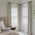 Photography of a neutral-toned bedroom interior featuring floor-to-ceiling beige pleated drapes on a black corner rod layered over white Roman shades a light wood bedside table with a book and candle and a textured knit throw blanket on the bed