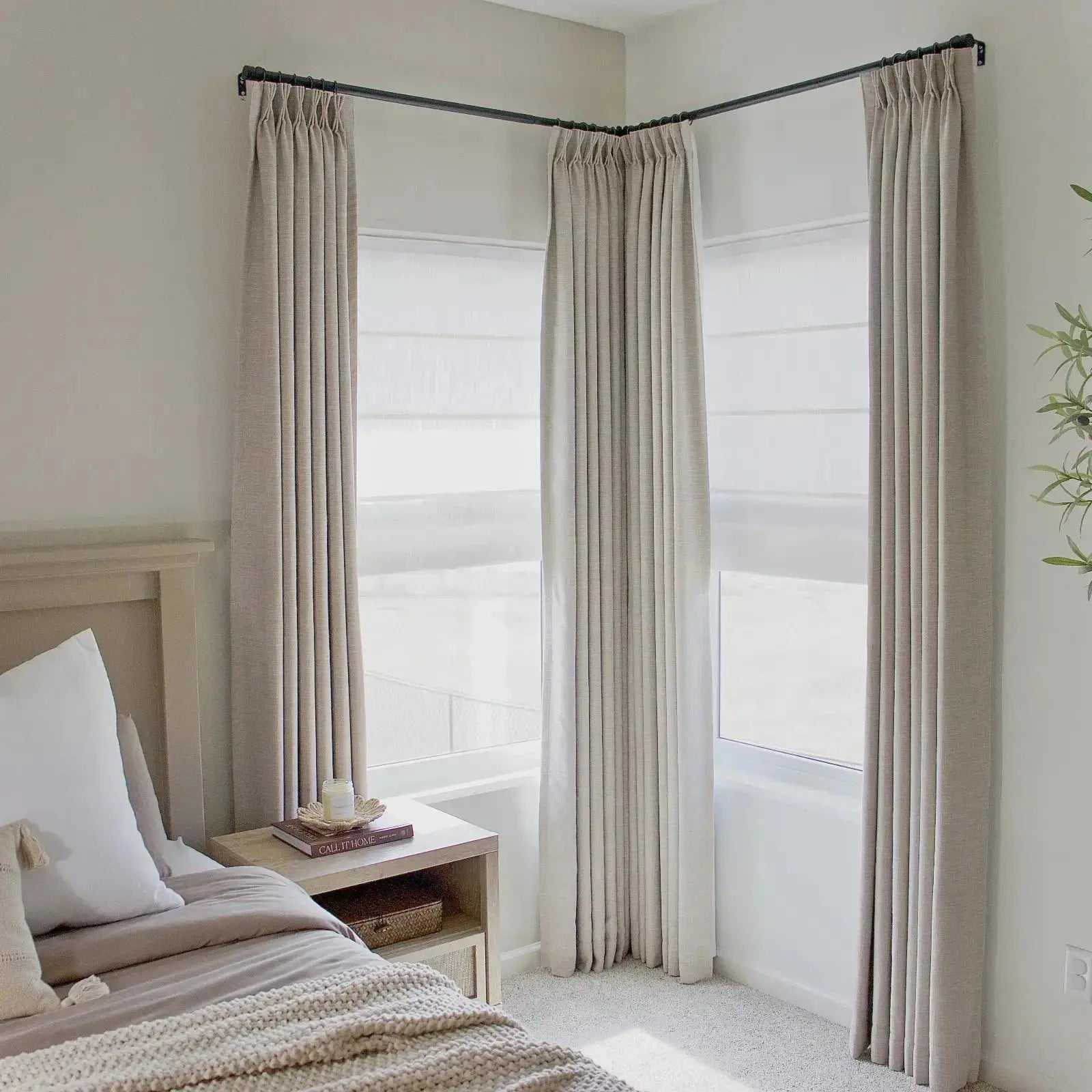 Photography of a neutral-toned bedroom interior featuring floor-to-ceiling beige pleated drapes on a black corner rod layered over white Roman shades a light wood bedside table with a book and candle and a textured knit throw blanket on the bed