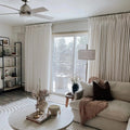A serene neutral living room interior features a Hunter ceiling fan, floor-to-ceiling white pleated curtains revealing a balcony with wintry trees, and a coffee table adorned with a gold bow candelabra holding white taper candles