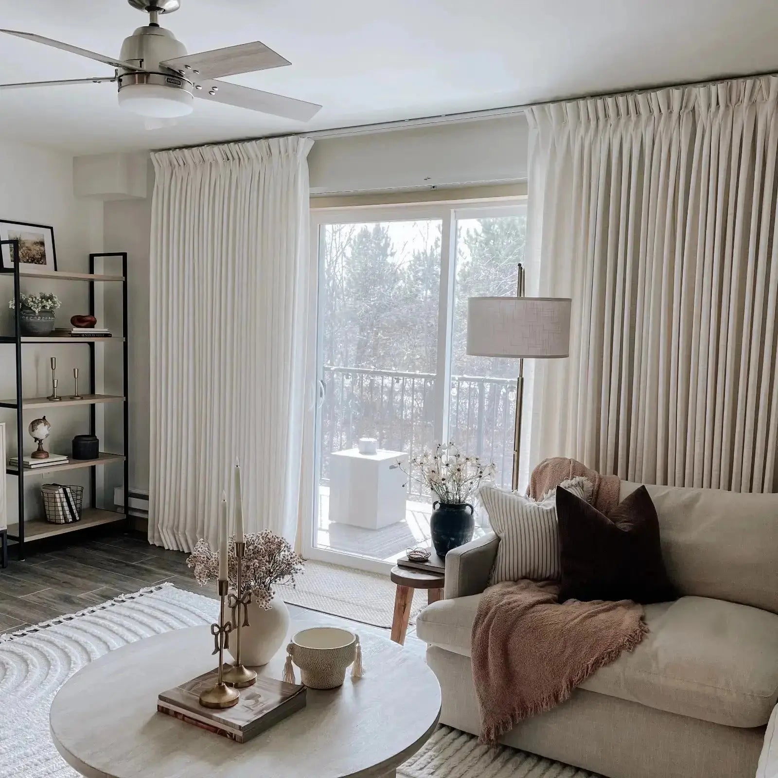 A serene neutral living room interior features a Hunter ceiling fan, floor-to-ceiling white pleated curtains revealing a balcony with wintry trees, and a coffee table adorned with a gold bow candelabra holding white taper candles
