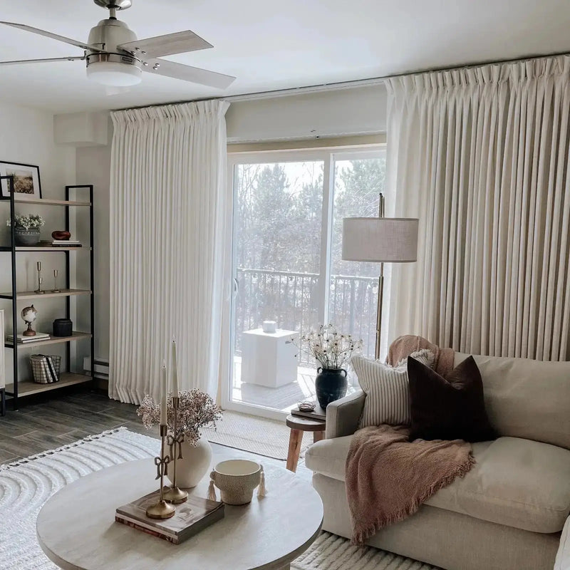 A serene neutral living room interior features a Hunter ceiling fan, floor-to-ceiling white pleated curtains revealing a balcony with wintry trees, and a coffee table adorned with a gold bow candelabra holding white taper candles