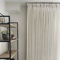 An indoor room featuring floor-to-ceiling cream pinch pleat curtains and a modern black metal and light wood bookshelf decorated with a dark grey pot of white flowers and brass candlesticks