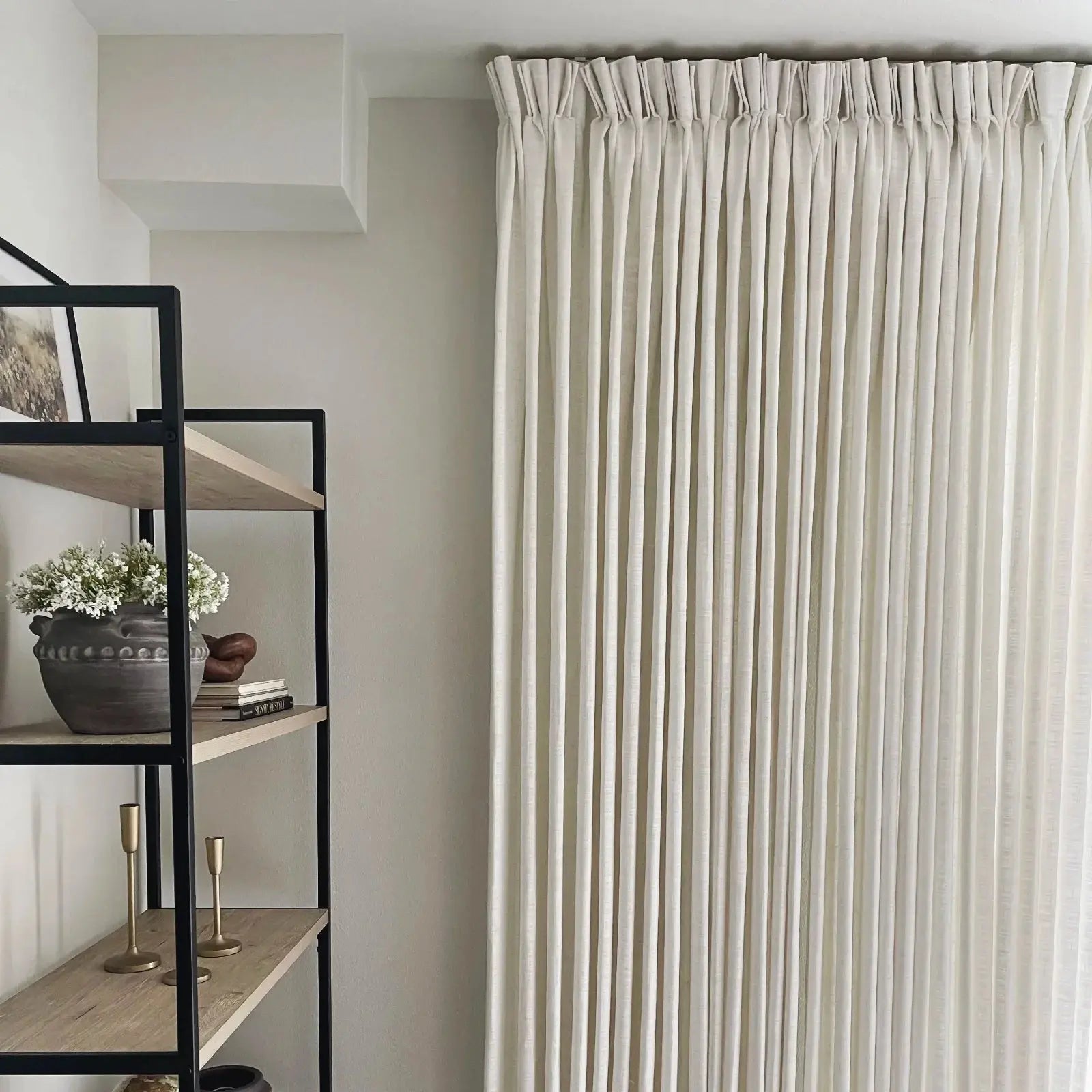 An indoor room featuring floor-to-ceiling cream pinch pleat curtains and a modern black metal and light wood bookshelf decorated with a dark grey pot of white flowers and brass candlesticks