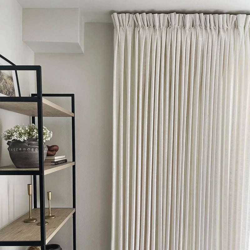 An indoor room featuring floor-to-ceiling cream pinch pleat curtains and a modern black metal and light wood bookshelf decorated with a dark grey pot of white flowers and brass candlesticks