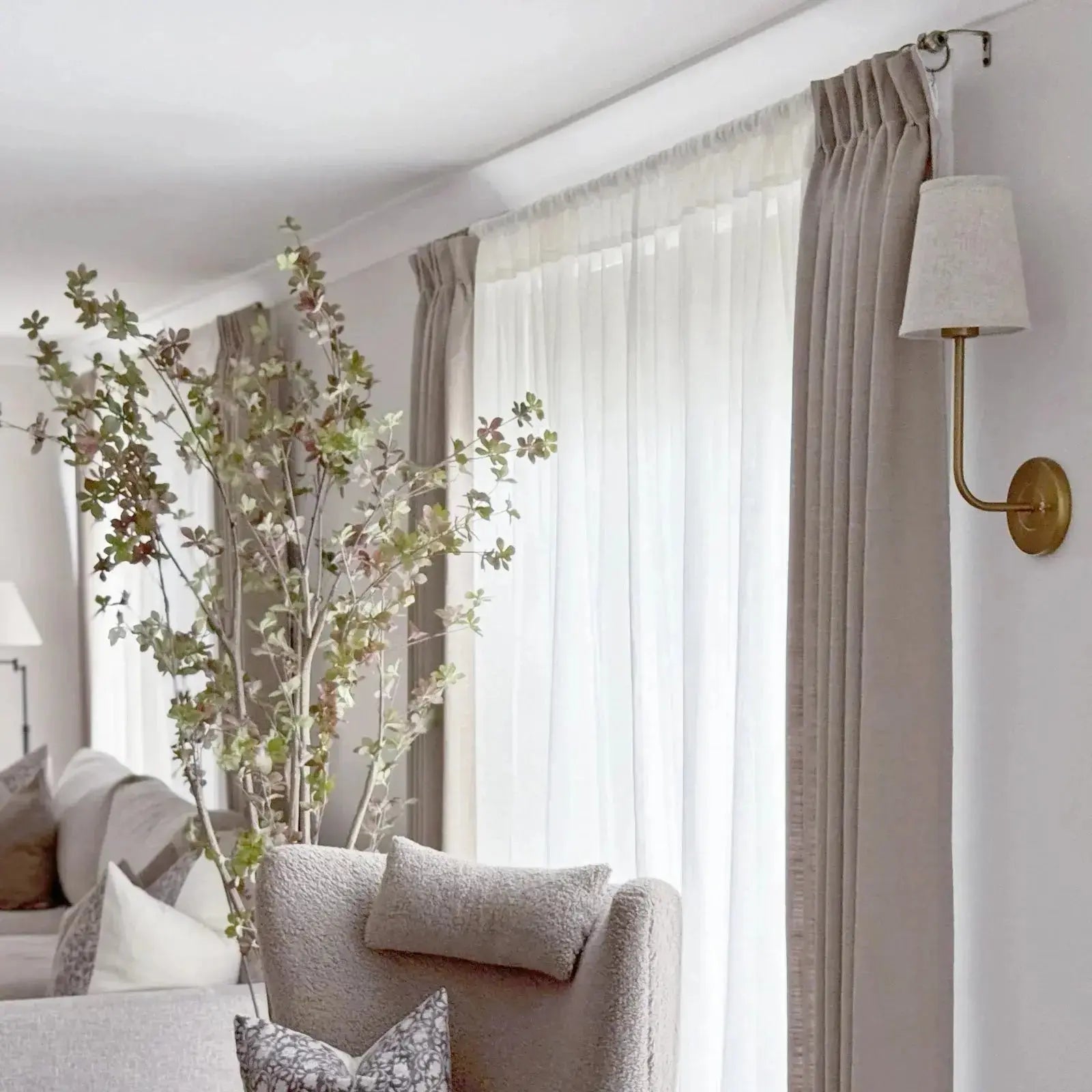 A cozy living room interior featuring tall decorative branches with small green leaves, a plush neutral armchair, floor-to-ceiling sheer white and beige curtains, and an elegant brass wall sconce