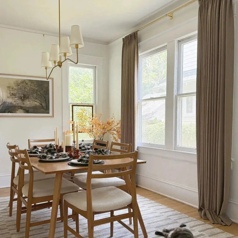 A cozy dining room featuring a wooden table set with fall decor including orange branches, tall candles, and plaid napkins, complemented by a brass chandelier, large windows with taupe drapes, and a grey tabby cat sleeping on a striped rug