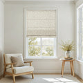 Photograph of a minimalist room with a window featuring a beige floral patterned Roman shade, a cream armchair with a wooden frame and a beige pillow, and a small round wooden side table holding a grey vase with green foliage