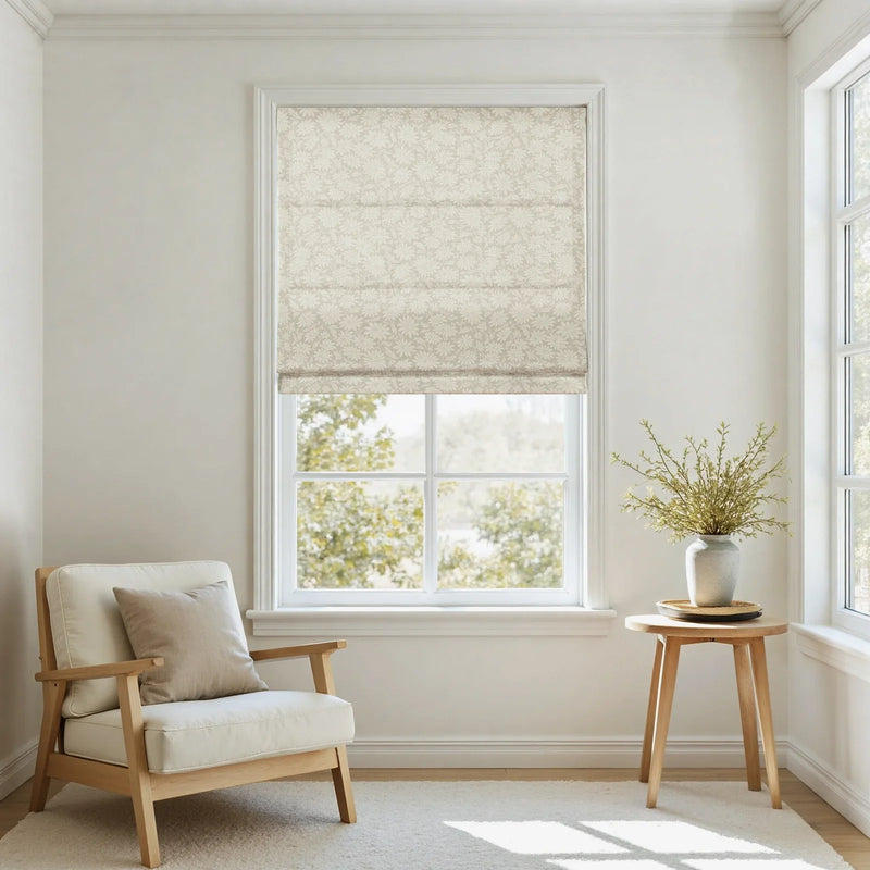 Photograph of a minimalist room with a window featuring a beige floral patterned Roman shade, a cream armchair with a wooden frame and a beige pillow, and a small round wooden side table holding a grey vase with green foliage