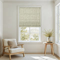 A bright and inviting room interior featuring a large window dressed with a light green leaf-patterned Roman blind, a cream upholstered armchair with a light wood frame, and a small round wooden side table holding a vase of green foliage