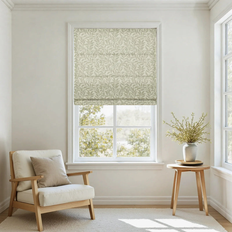 A bright and inviting room interior featuring a large window dressed with a light green leaf-patterned Roman blind, a cream upholstered armchair with a light wood frame, and a small round wooden side table holding a vase of green foliage