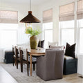 A cozy modern farmhouse dining room with white shiplap walls, centered around an oval wooden table under a dark brown woven pendant light, adorned with a tall distressed green vase of greenery and flanked by upholstered seating and multiple windows dressed in light linen Roman shades